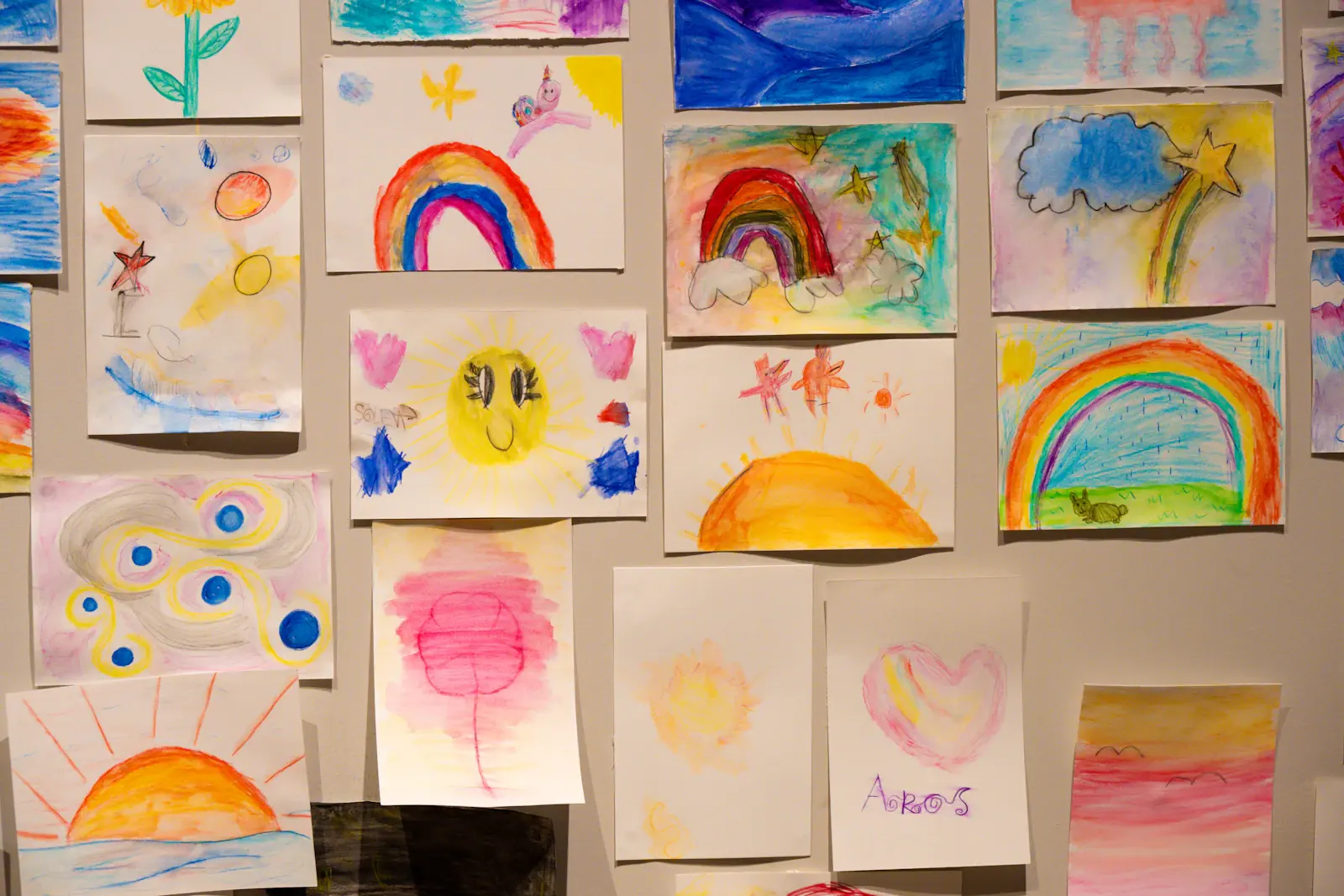 Children's drawings pinned side by side on a wall: rainbows, suns, flowers and a heart.